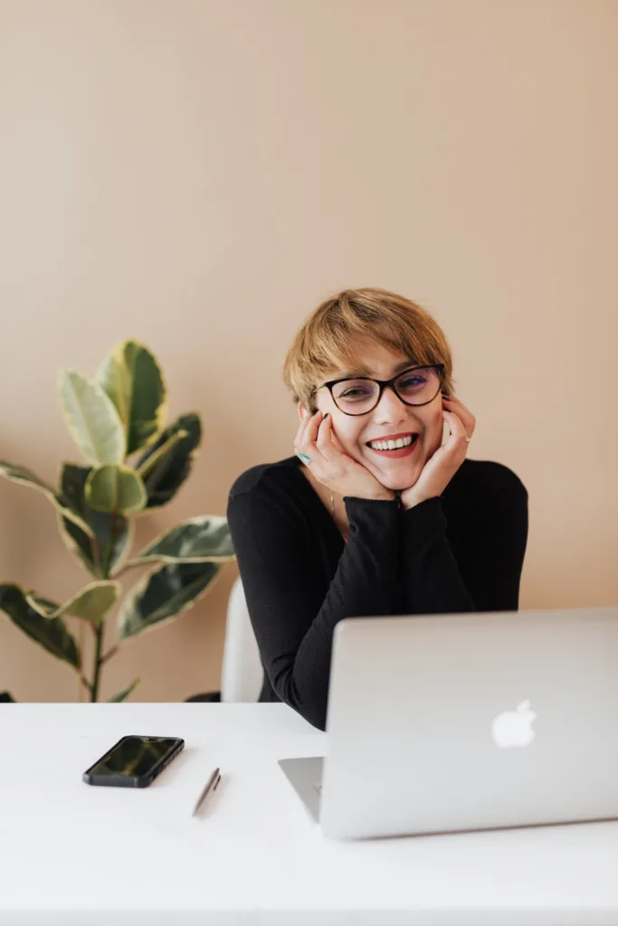 ADHD Careers that don't suck for ADHD adults. Image of a girl with short blonde hair sitting behind a mac book at a clean desk, holding her face in her hand, very happy and smiling. 