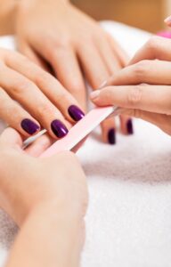 A close-up view shows a pair of hands using a pink nail file to down the nails on another pair of hands with purple polish.