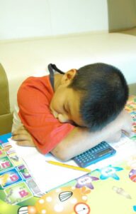 A boy with his head down on a desk asleep. There is a calculator, paper, and a pencil underneath his arms on the desk.