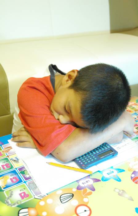 A boy with his head down on a desk asleep. There is a calculator, paper, and a pencil underneath his arms on the desk.