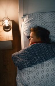 A little girl tucked into bed beneath a white and blue comforter. She sleeps with her nightstand lamp on.