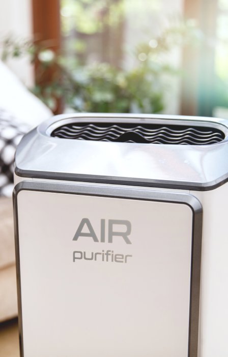 A white air purifier contains the words "air purifier" on the front. A woman sits on a cream couch looking at a book.