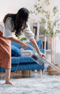 A woman vacuums a plush, light gray rug in front of a blue couch. Sunlight from a large window fills the living room.