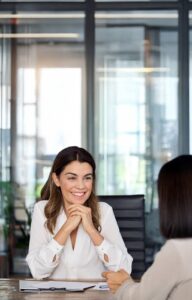 Two women sit in a conference room at a table across from each other. One woman has her hands under her chin, listening.