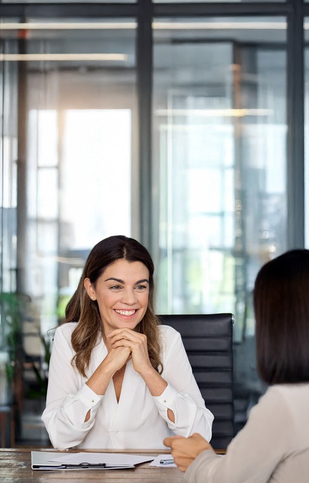 Two women sit in a conference room at a table across from each other. One woman has her hands under her chin, listening.