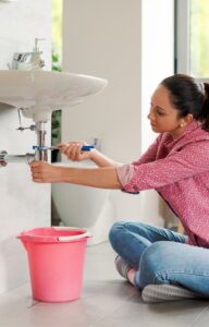A woman wearing a floral shirt sits on the bathroom floor using a wrench to twist an exposed pipe under the sink.