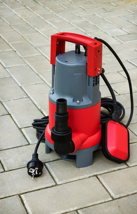 A gray and red sump pump sits on a gray stone floor and is unplugged. It has a long, black cord.