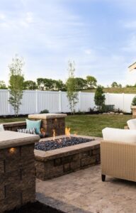 A stone patio in a backyard surrounded by a white fence. There is a fire pit and pergola on the patio.