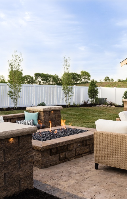 A stone patio in a backyard surrounded by a white fence. There is a fire pit and pergola on the patio.