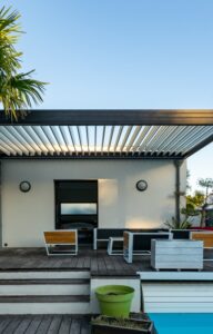 A black pergola sits on a wooden two-level deck with chairs and a table underneath it. A palm tree is next to the pergola.