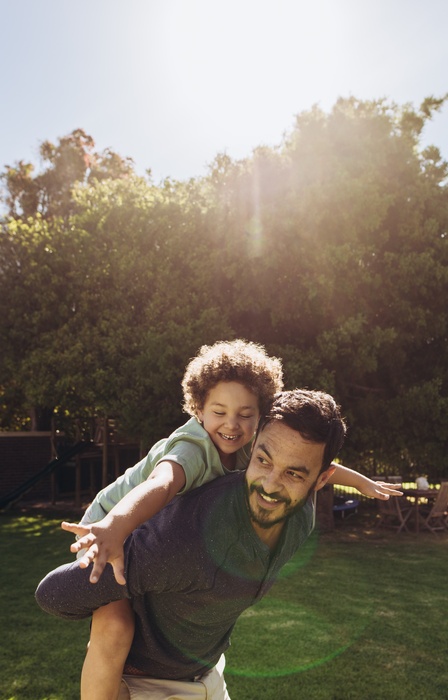 A young boy sits on his dad's shoulders while he runs around the park. The sun shines through the trees.