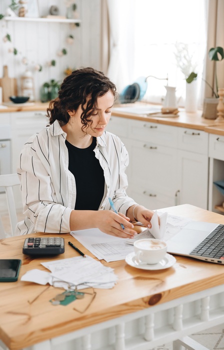 A woman sits at a kitchen countertop with a laptop, paperwork, a calculator, and coffee while budgeting.