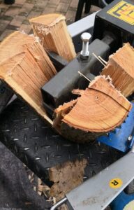 A close-up view shows a black, electric log splitter splitting a large piece of wood into four quarters.