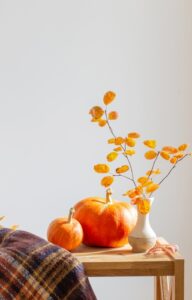 A wooden side table features two orange pumpkins, and a white vase holds a twig with orange leaves, placed next to a couch.