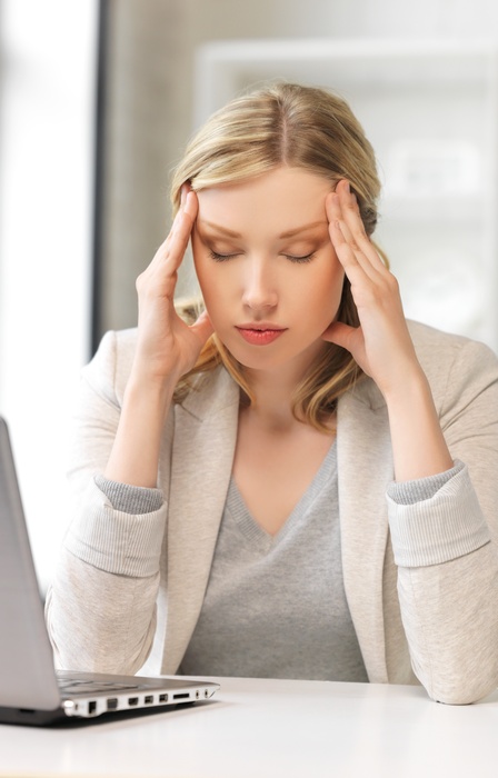 A woman sits at a white desk, closing her eyes and massaging her temple. An open laptop sits on the desk.
