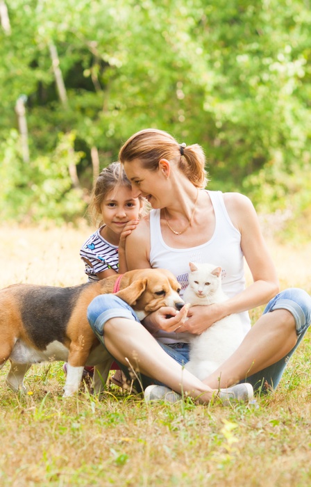 A woman and young girl sit in a grassy field with a white cat and a brown-and-white dog in their arms.