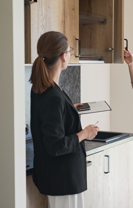 A young blonde woman opening a kitchen cabinet and examining the interior in front of a woman carrying a clipboard.