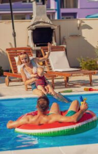 A woman sits on the edge of a pool holding a baby as a toddler sprays them with a water gun. A man lies on a float in the pool.