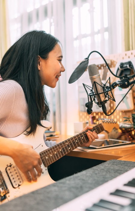 A woman holding a guitar and sitting in front of a keyboard and digital technology leans into a studio microphone.