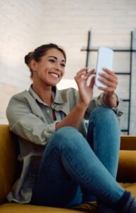 A woman sits on a mustard-colored couch. She holds her smartphone in front of her face and smiles at it.