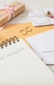 A table has a spiral notebook with a page stating "Wedding Checklist." There are white envelopes, gold scissors, and a manila envelope as well.