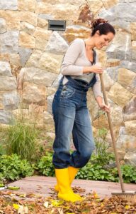 A smiling woman in overalls and bright yellow rain boots is sweeping leaves on a patio deck into a pile.