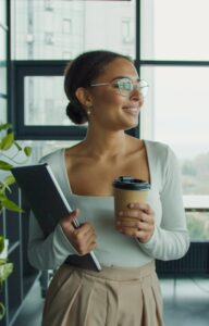 A young woman wearing glasses and a white shirt holds a coffee cup and laptop while standing in front of a large window.