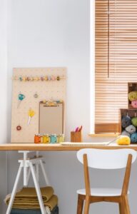An organized crafting room contains a white chair, a wooden desk, crates with colorful yarn, and a pegboard with scissors.