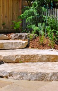 Four oversized stone steps lead to a light-colored wooden gate. Mulch, small rocks, and flowers line the steps.