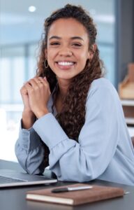A smiling, long-haired woman in a blue blouse sitting at a desk with an open laptop and a small brown notebook.