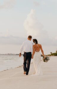 A woman in a white dress walks along a sandy beach near the water, holding a man's hand and a bouquet.
