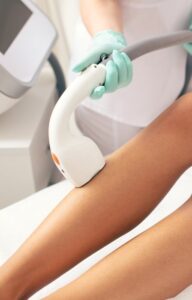 A woman with a red manicure, wearing a short white robe, undergoes laser hair removal on her right leg.