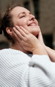 A woman in a white sweater holds her hands to her cheeks, tilts her head back, and smiles with her eyes closed.