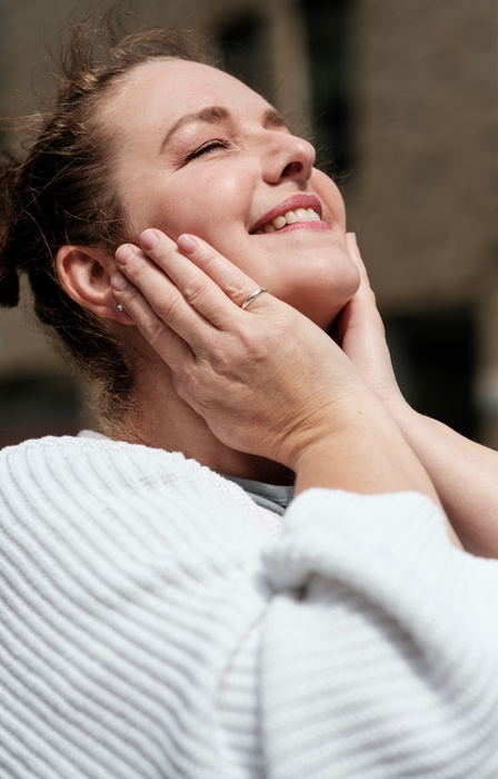 A woman in a white sweater holds her hands to her cheeks, tilts her head back, and smiles with her eyes closed.