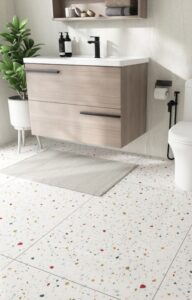 A modern bathroom features a white tile with polka-dots, a white toilet, a floating vanity that is brown, and a potted plant.