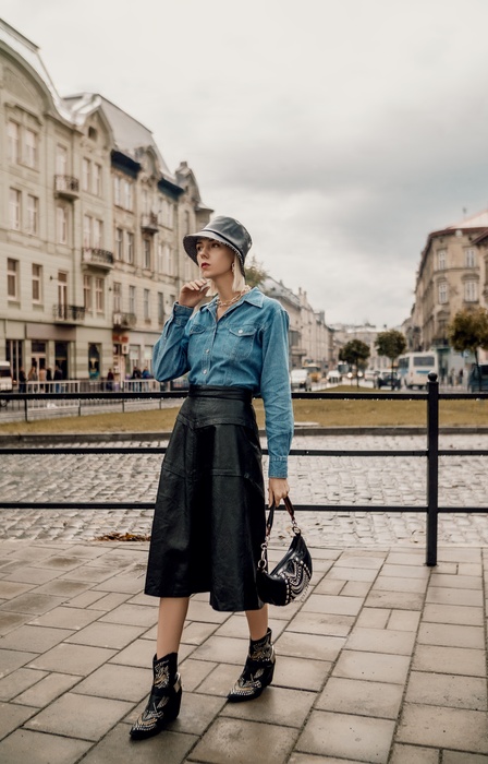 A woman in a bucket hat, denim shirt, long skirt, and cowgirl boots walks on a stone-paved city street carrying a purse.