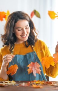 A woman smiles as she creates decorations with autumn leaves on a string. Other craft projects are in the room.