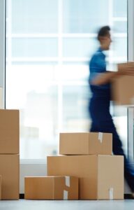 An employee carrying a box and walking next to big windows. Nine more boxes of various sizes are on the floor.