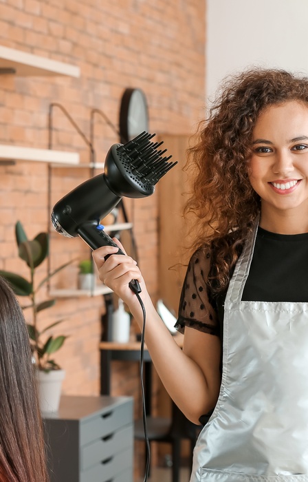 A woman looking at the camera with multiple hair care products in her hands. There is another woman in the chair next to her.