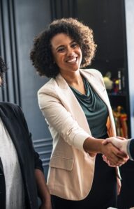 Two professional Black women, both smiling, one shakes hands with a man during a business meeting in a modern office.