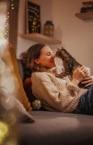 A Caucasian woman sits on a sofa cuddling a small cat in a dimly lit room, illuminated by a lamp beside them.