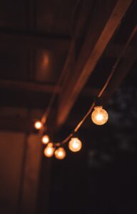 A close-up of string lights hung from wooden beams outside at night. The bulbs put off a warm, orange glow.