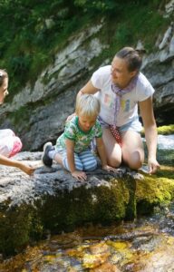 Woman and two children kneeling on mossy rocks beside a shallow forest stream, exploring nature together.
