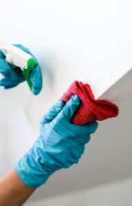 A close-up view shows a person wearing blue gloves using a red cloth to wipe down a wall after spraying liquid on it.