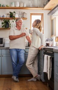 An older man and woman are standing in a kitchen; both are holding coffee mugs and engaged in conversation.
