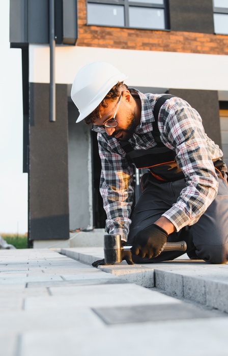 A worker using a small mallet to install concrete driveway pavers outside a home during the daytime.