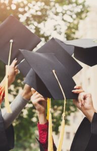 A group of arms of new graduates holding up their caps together. They are all wearing graduation gowns.
