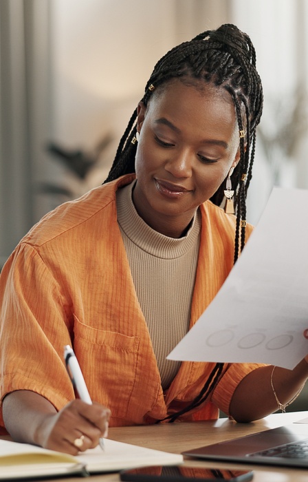 Woman with braided hair writes in notebook while holding paper, seated at desk with laptop and phone in bright workspace.