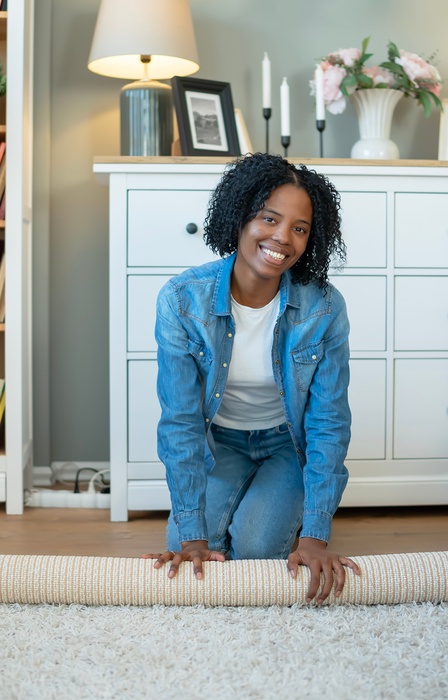 A woman unrolling a carpet across the floor in a living room, with furniture and lamps visible in the background.