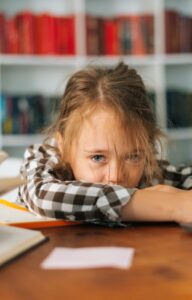 A frustrated and worn out girl rests her head on her arms at a cluttered desk with books and a laptop in a classroom.